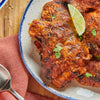 Grilled chicken thighs, richly coated in a reddish-orange spice rub, are presented on a speckled plate with a lime wedge and cilantro, showcasing a dish prepared with Lawry'S Casero Chili Powder.