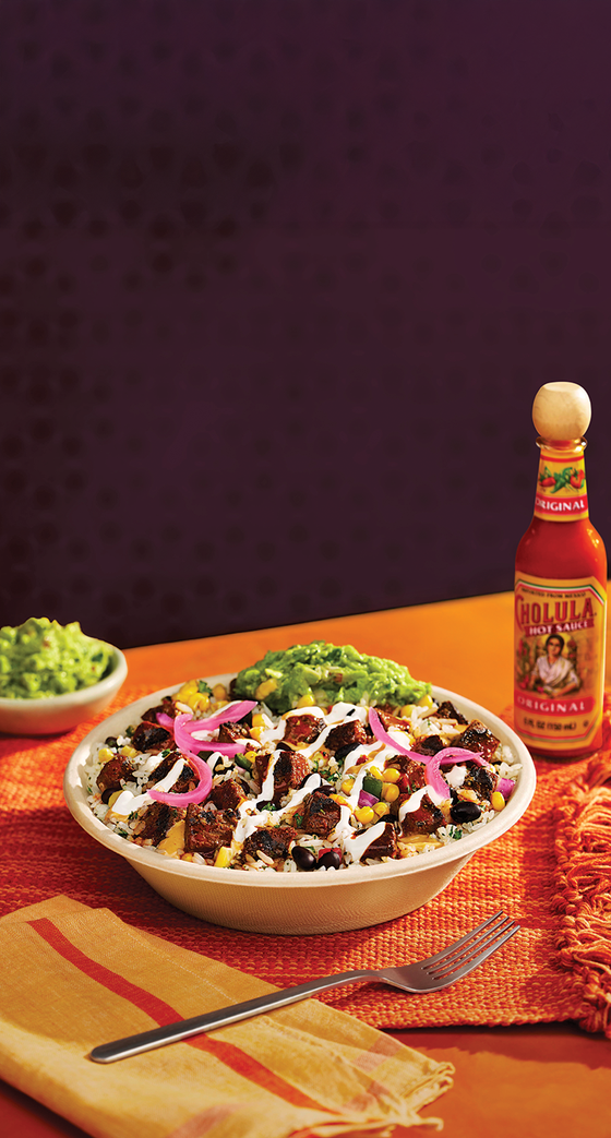 Bowl of taco salad with guacamole and a bottle of hot sauce on an orange tablecloth.