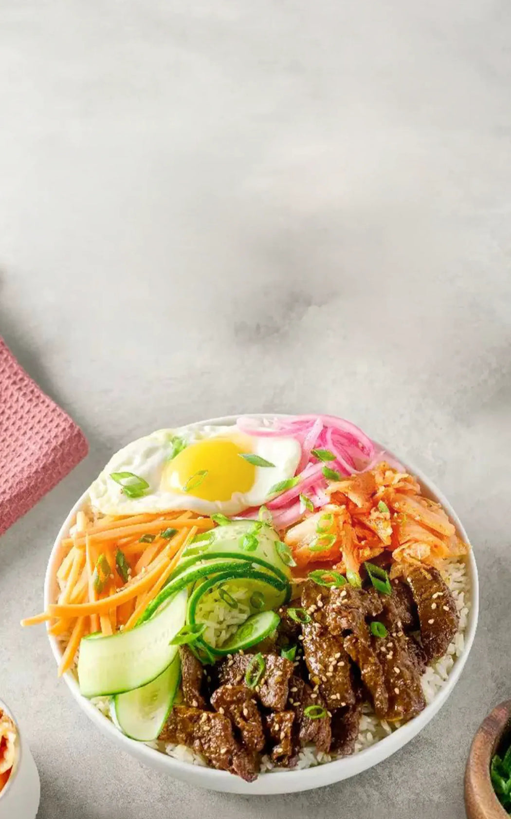 Bowl of Korean bibimbap with beef, vegetables, and a sunny-side-up egg on a light background.