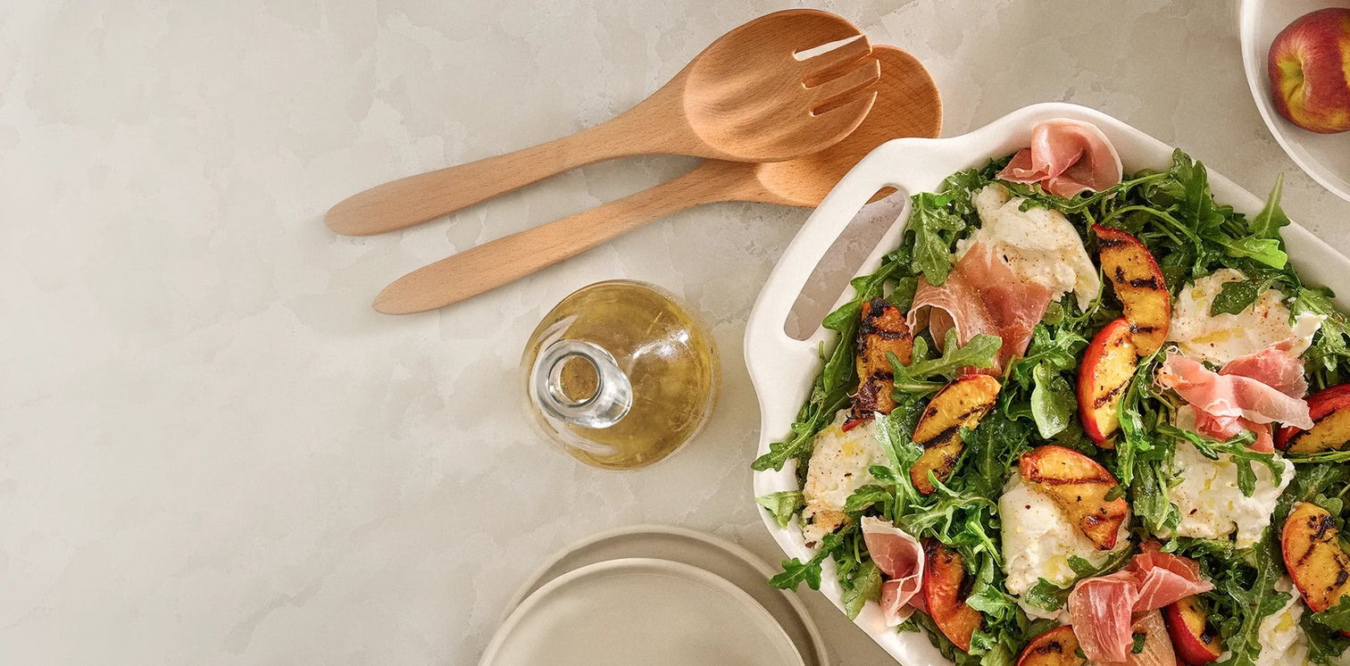 Salad with prosciutto and peaches on a table with wooden utensils and a glass of white wine.