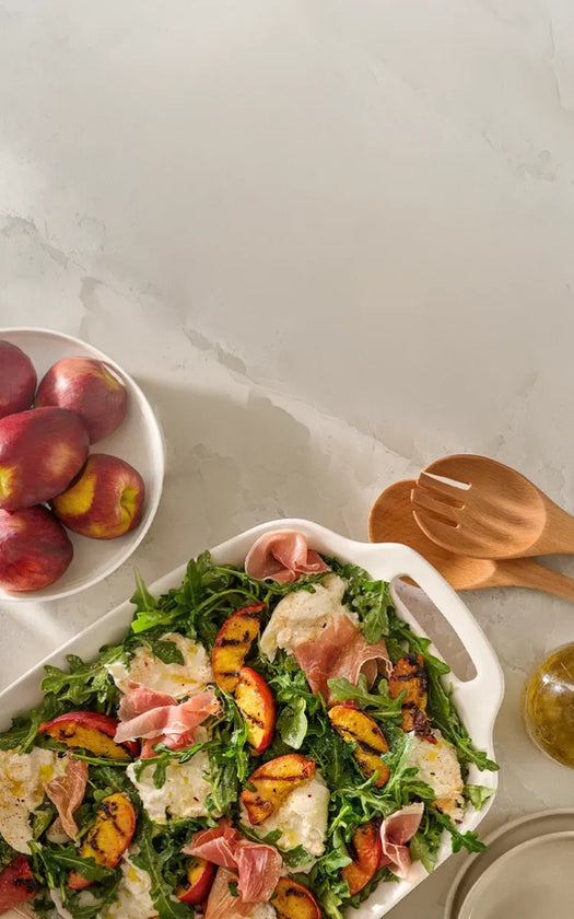 Salad with prosciutto and peaches on a white plate with a bowl of peaches and wooden utensils in the background.