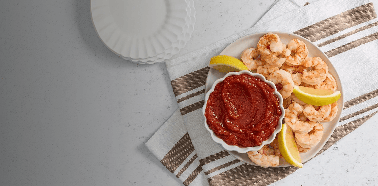 Platter of shrimp with a bowl of red sauce and lemon wedges on a striped cloth.