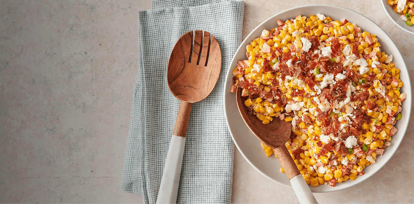 Corn salad with bacon and cheese in a white bowl on a gray surface with a wooden spoon.