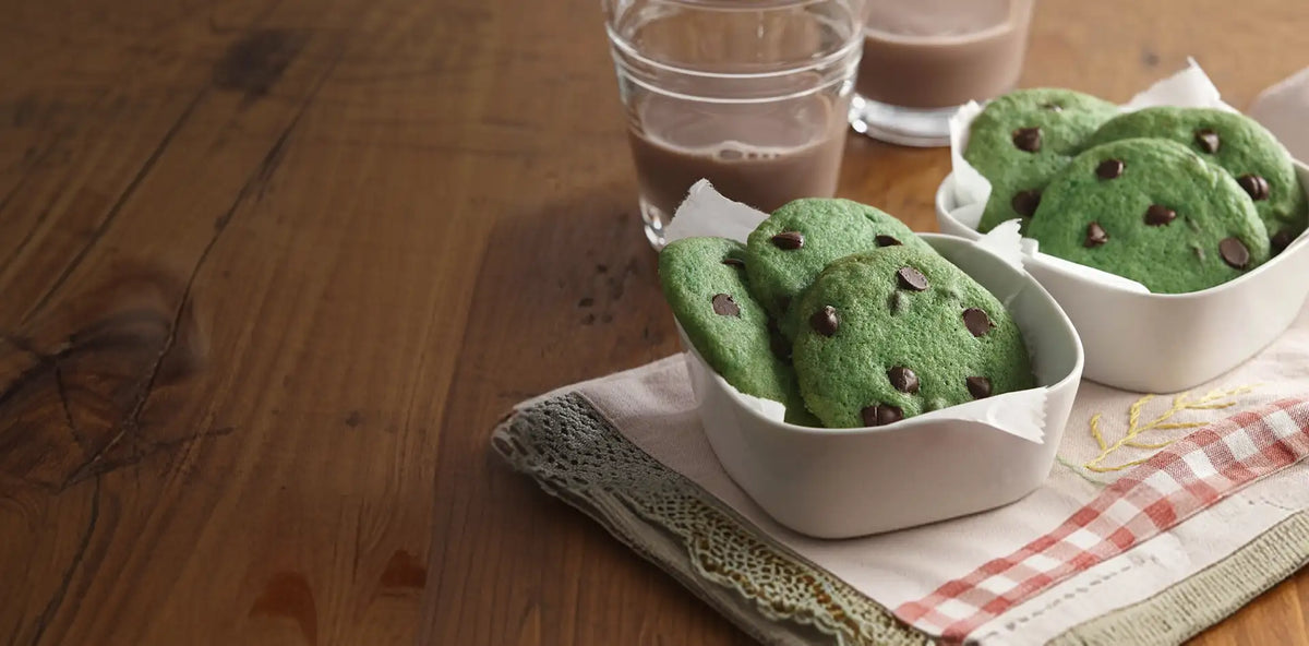 Green cookies with chocolate chips in a white bowl on a wooden table with a glass of milk.