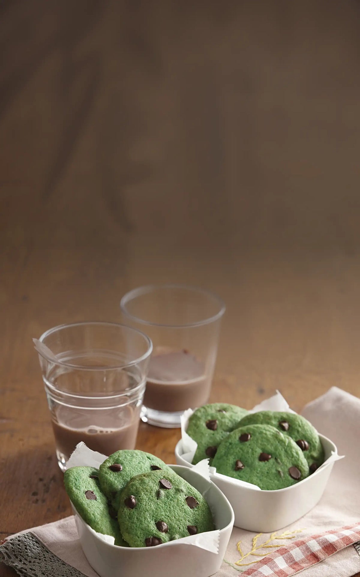 Green cookies with chocolate chips in white bowls on a wooden table with two glasses of milk.