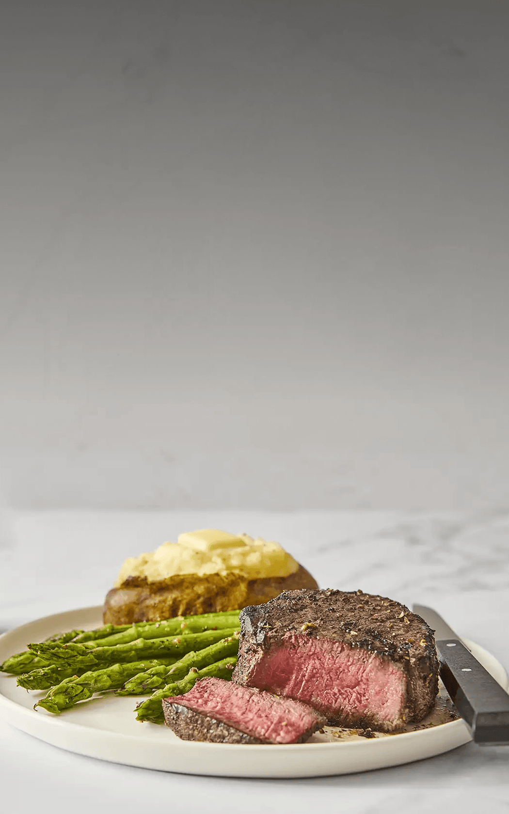 Steak with asparagus and baked potato on a plate with a knife, against a light gray background