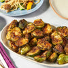 A plate of roasted Brussels sprouts is displayed alongside a plate of pasta with vegetables.