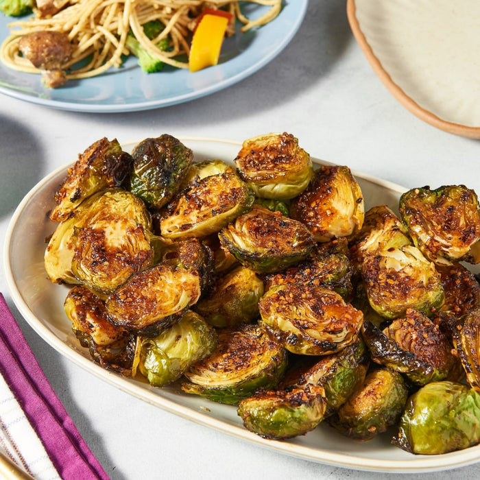 A plate of roasted Brussels sprouts is displayed alongside a plate of pasta with vegetables.