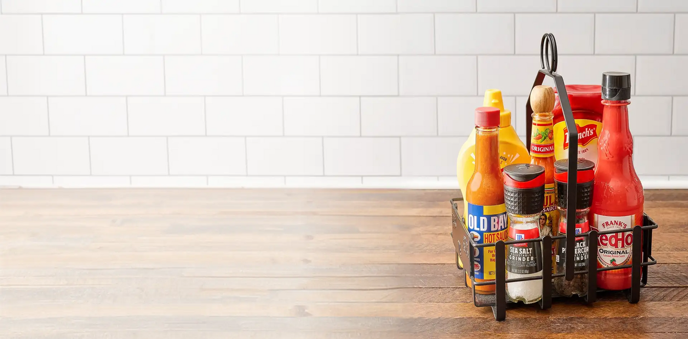 Assorted sauce bottles in a metal caddy on a wooden surface with a white tiled wall background.