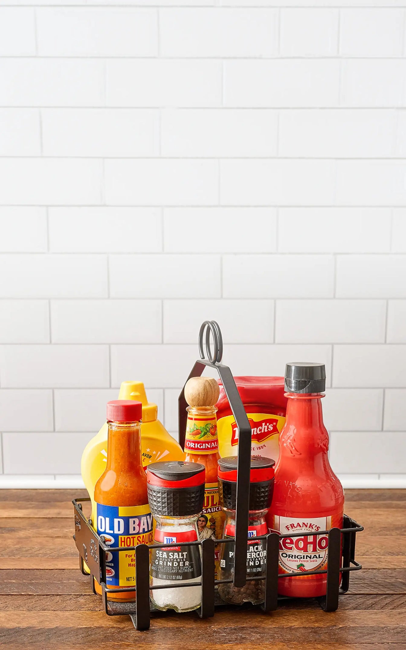 Assorted sauce bottles in a metal caddy on a wooden surface with a white tiled wall background.