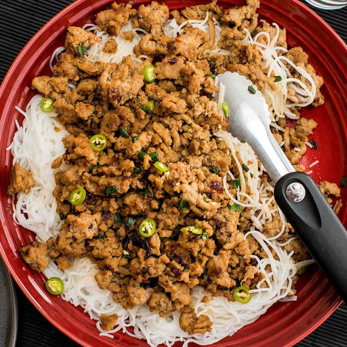 A red bowl of rice noodles topped with ground pork, green onions, and chili peppers.