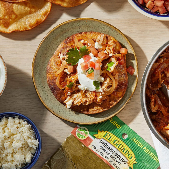 A plate of El Guapo brand ground oregano with a tostada topped with chicken, onions, tomatoes, and sour cream.