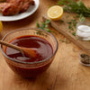 A bowl of barbecue sauce with a wooden spoon in it, next to a plate of chicken and a cutting board with lemons, herbs, and spices.