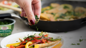 Person garnishing a dish with herbs over a plate of food, with a pan and ingredients in the background.