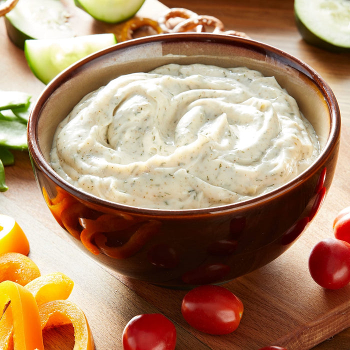 A bowl of creamy dip with dill and parsley sits on a wooden cutting board with fresh vegetables.