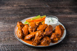 A plate of buffalo wing wings in a rich orange-brown sauce, served with celery sticks, carrot sticks, and a small glass of blue cheese dip on a dark rustic wooden table.