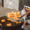 A metal strainer filled with deep-fried dough is being lifted out of a pot of hot oil.