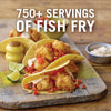 A close-up of a wooden cutting board with three fish tacos, a yellow salt shaker, and a bowl of dipping sauce.