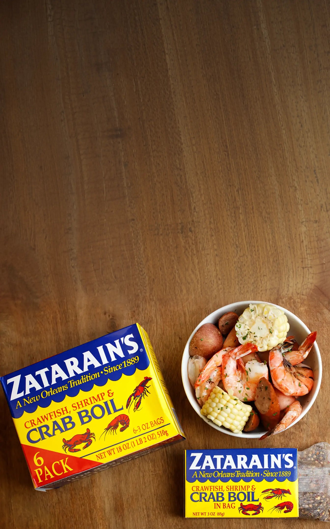 Zatarain's Crawfish, Shrimp & Crab Boil 6-pack and single bag next to a bowl of boiled shrimp, corn, and potatoes