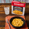 A box of Zatarains Cajun-Style Mac &amp; Cheese sits next to a glass of water and a bowl of the prepared mac and cheese with a fork.