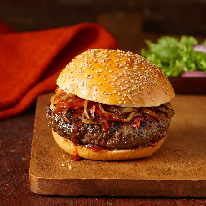 A juicy burger with caramelized onions and a sesame seed bun sits on a wooden cutting board.