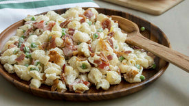 Pasta salad with bacon and peas on a wooden plate with a wooden spoon.
