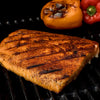 A seasoned salmon fillet with grill marks rests on a dark grill grate, accompanied by grilled bell peppers in the background, suggesting the use of Mccormick Grill Mates Barbecue Seasoning.