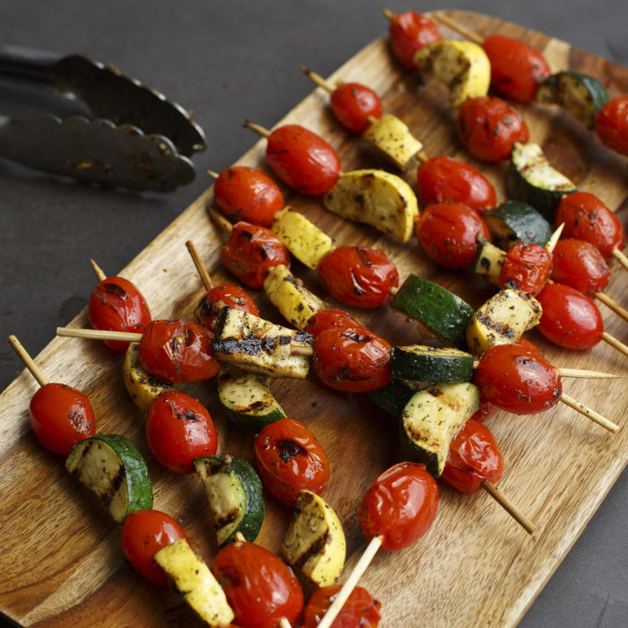 Grilled vegetable skewers with tomatoes and zucchini are arranged on a wooden board next to a pair of tongs.