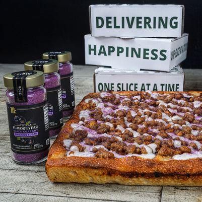 A rectangular slice of pizza topped with purple berries, brown cereal-like pieces, and white drizzle, displayed on a rustic wood surface next to several glass jars of berry-flavored syrup and stacked white delivery boxes with green text.