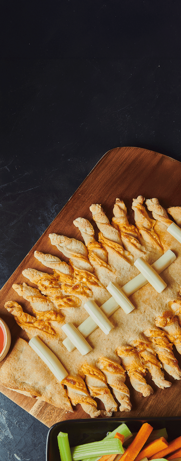 A wooden serving board arranged with twisted tortilla chips drizzled with orange sauce, paired with white cheese sticks, celery, carrot sticks, and a small dipping sauce on a dark background.