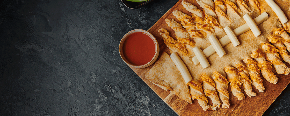 A wooden board filled with flatbread topped with twisted cheese sticks and several small bowls of red dipping sauce, photographed on a dark textured surface with negative space on the left.