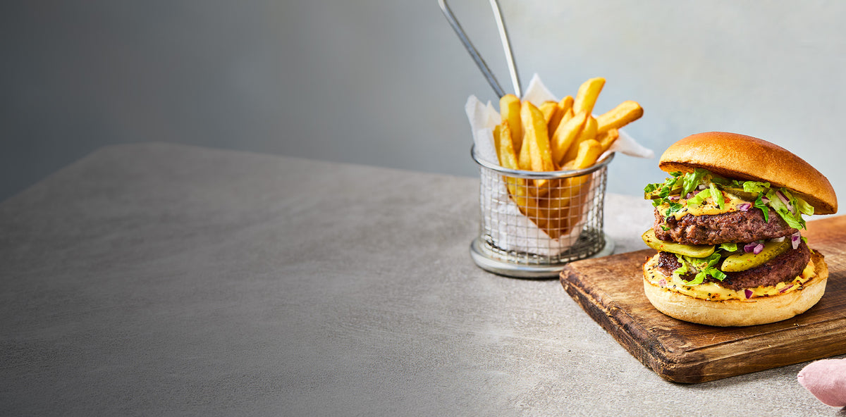 A gourmet burger with a beef patty, lettuce, and pickles on a wooden board, served with a side of french fries in a wire basket on a gray tabletop.
