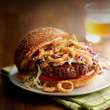 Close-up of a burger with coleslaw, crispy onions, tomato, and creamy sauce on a sesame seed bun, served on a plate.