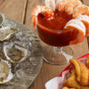 A plate of oysters and a glass of shrimp cocktail with a basket of fried food in the background.