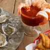 A plate of oysters and a glass of shrimp cocktail with a basket of fried food in the background.