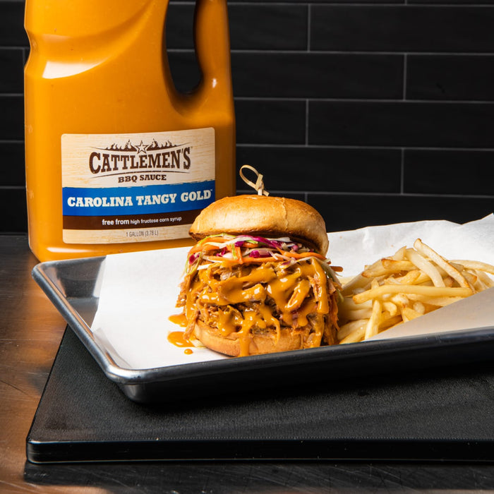 A pulled pork sandwich with coleslaw and a side of fries, next to a bottle of Cattlemens Carolina Tangy Gold BBQ sauce.