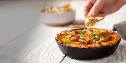 Hand dipping a chip into a bowl of nachos with cheeseburger dip on a wooden table.