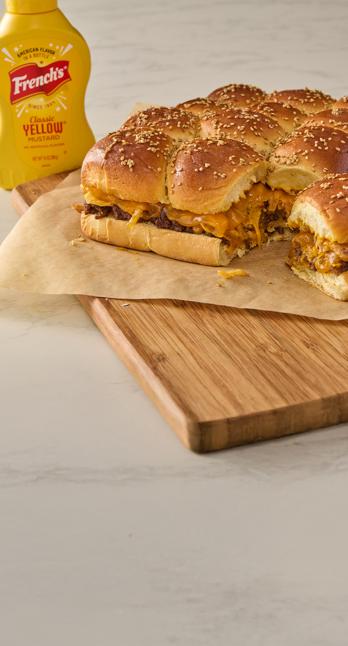 Burgers on buns with cheese on a wooden cutting board next to a bottle of French's yellow mustard.