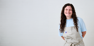 Chef Morgan smiling in a light blue shirt and beige apron, standing against a clean white tiled wall background.