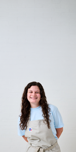 Chef Morgan smiling in a beige apron and light blue shirt, standing against a white tiled wall with ample negative space above.
