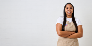Chef Taylor smiling with arms crossed, wearing a beige apron and white shirt, standing against a white tiled wall with negative space on the left.