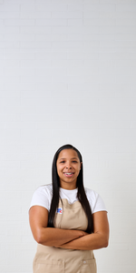 Chef Taylor smiling with arms crossed in a beige apron and white shirt, standing against a white tiled wall with ample space above.