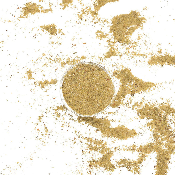 A bowl of dried herbs and spices sits on a white background with some of the spices scattered around it.