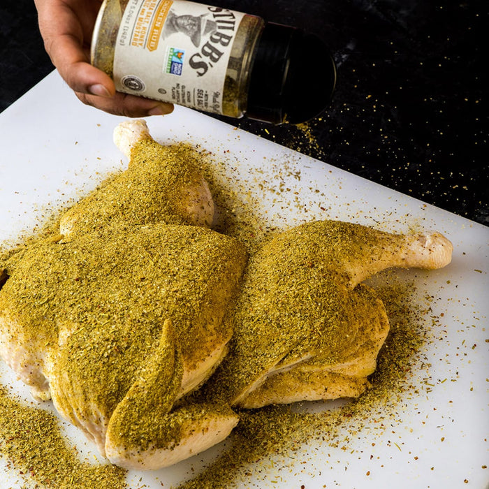 A hand is sprinkling a chicken with BBs Chicken Rub seasoning.