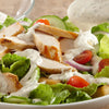 A close-up of a salad with grilled chicken, cucumbers, tomatoes, red onion, and a creamy dressing.