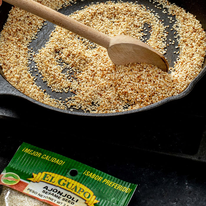 A close up of a cast iron skillet with sesame seeds being toasted with a wooden spoon, and a bag of El Guapo sesame seeds.