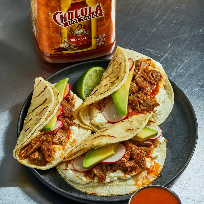 Three tacos filled with meat, avocado, radish, and sour cream are on a black plate with a bowl of Cholula hot sauce.
