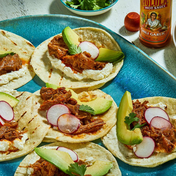 A plate of tacos topped with shredded beef, avocado, radish, and cilantro, with a bottle of Cholula Chili Garlic Hot Sauce in the background.