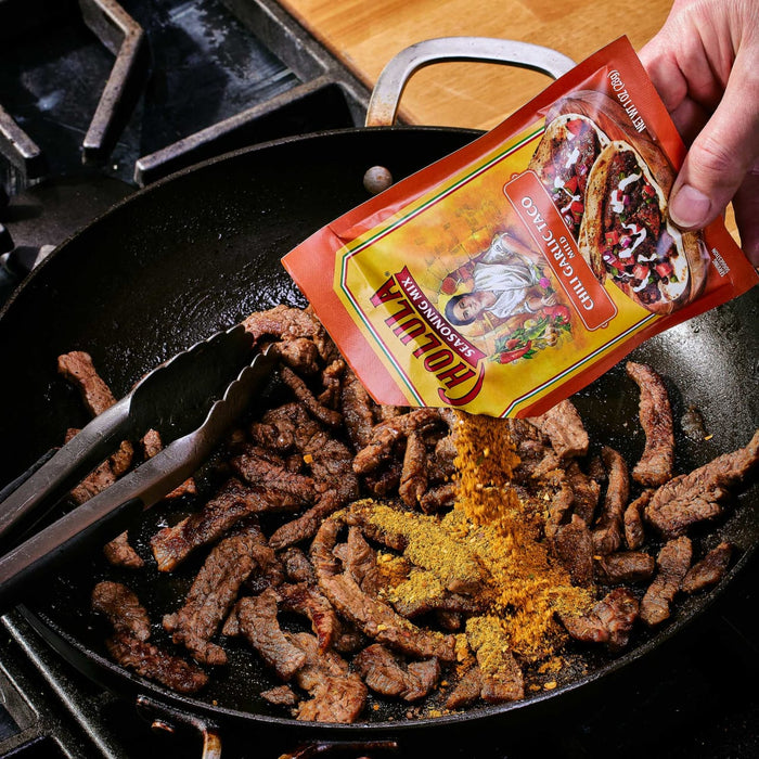 A hand sprinkles Cholula Chili Garlic Taco seasoning on a pan of cooked beef strips.