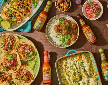 Assorted Mexican dishes including tacos, enchiladas, and bowls of rice and beans on a wooden table with hot sauce bottles.